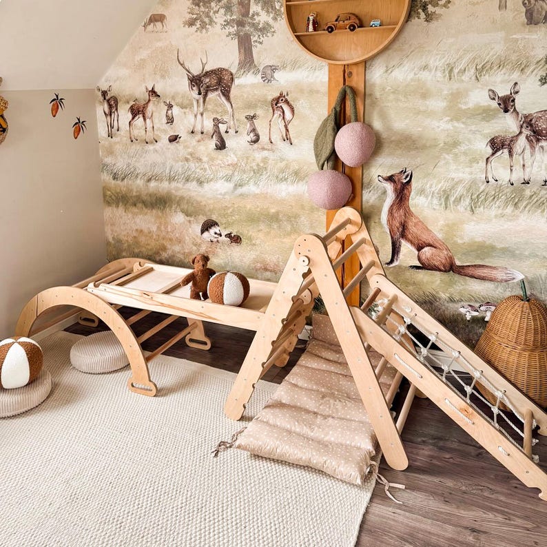 May include: A children's playroom with a wooden climbing arch, a slide, and a table. The room features a forest-themed mural with deer and a fox. Decorative elements include a round shelf, hanging pink spheres, and a woven basket.