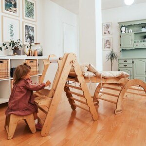 May include: A wooden children's play set featuring a climbing frame, a ramp with a cushion, a climbing arch, and an easel with a paper roll. The set is made of light-colored wood and is designed for indoor play. A child is drawing on the easel.
