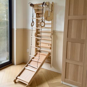 K&ouml;nnte beinhalten: Ein helles, an der Wand montiertes Kletterger&uuml;st aus Holz mit einer vertikalen Leiter, einer abnehmbaren schr&auml;gen Rutsche, einer Strickleiter mit Holzsprossen, zwei h&auml;ngenden Turnringen und einer runden Holzscheibe an einem dicken Seil.