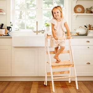 May include: A wooden and white step stool with a child standing on it in a kitchen. The step stool has a platform and a bar for the child to hold onto. The child is wearing a light-colored outfit.