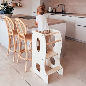 May include: A white wooden kitchen step stool with a natural wood step and handrails. The step stool has rounded cutouts and is designed to help a child reach countertops. A child is standing on the step stool.