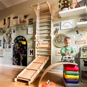 Könnte beinhalten: Ein hölzerner Kletterrahmen mit Leiter, Seil und Rutsche. Ein Kind spielt mit einem Spielzeugauto auf einem Regenbogen-Balanceboard. Der Raum hat dekorative Schilder, darunter "COOL KIDS CLUB" und "WELCOME TO MY CRIB".