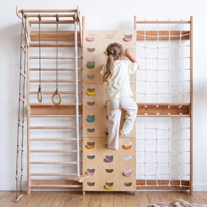 May include: A wooden indoor climbing gym featuring a wall ladder, a rope ladder, hanging rings, a climbing rope with a disc seat, and a central climbing wall with colourful handholds. The structure is made of light-coloured wood with white rope.