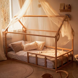 May include: A wooden house-shaped floor bed frame with a low-profile design and safety rails. The frame is draped with a sheer, light-colored fabric canopy. The bed is dressed with a white mattress, a brown pillow, and a textured cream pillow.