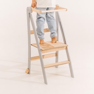 May include: A gray and natural wood learning tower with a safety bar. The tower has a step and a platform for a child to stand on. The frame is gray, and the steps and platform are natural wood.