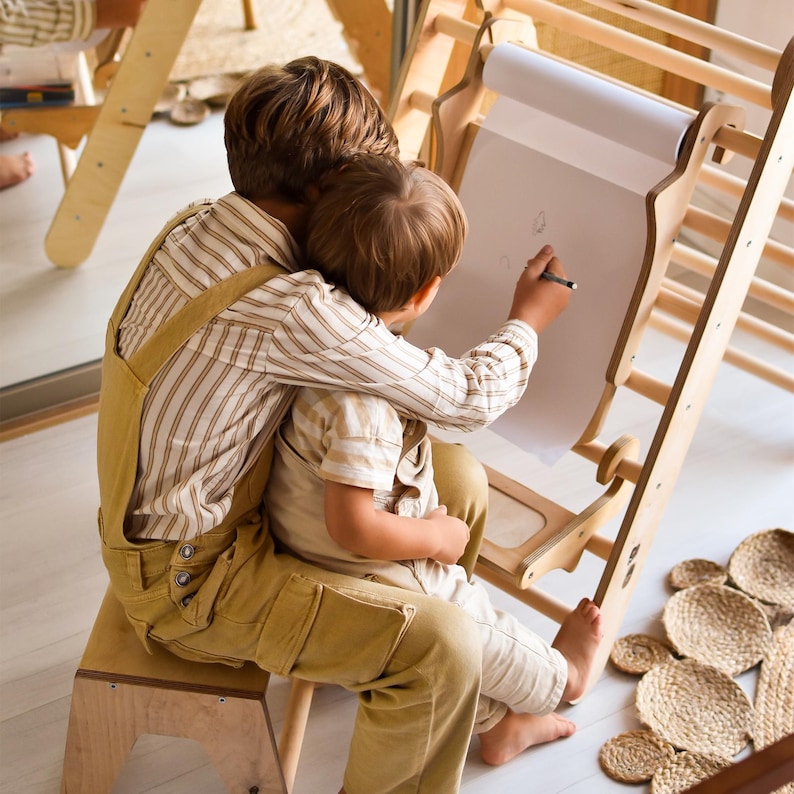May include: Two children are seated, one drawing on a white paper roll attached to a wooden easel. The children are wearing matching mustard-colored overalls and striped shirts. A wooden stool and a woven mat are also visible.
