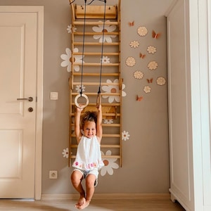 Könnte beinhalten: Ein hölzerner Kletterrahmen mit Hängeringen. Der Rahmen ist an einer grauen Wand montiert, die mit weißen Gänseblümchen- und Schmetterlingsaufklebern verziert ist. Ein Kind hängt an den Ringen. Eine weiße Tür und ein Schrank sind sichtbar.