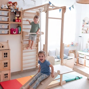 Könnte beinhalten: Ein hölzernes Klettergerüst und Rutschbahn-Set in einem Kinderspielzimmer. Ein Junge klettert die Leiter hoch, und ein Mädchen rutscht die Rutsche hinunter. Der Raum hat bunte Matten, Spielzeug und dekorative Elemente.
