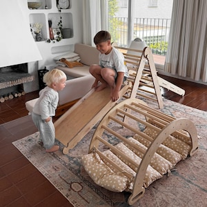 May include: Wooden climbing arch, slide, and climbing triangle set. The arch has a series of wooden rungs and is paired with star-patterned cushions. The slide and triangle are also made of wood. Two children are playing on the set in a living room.