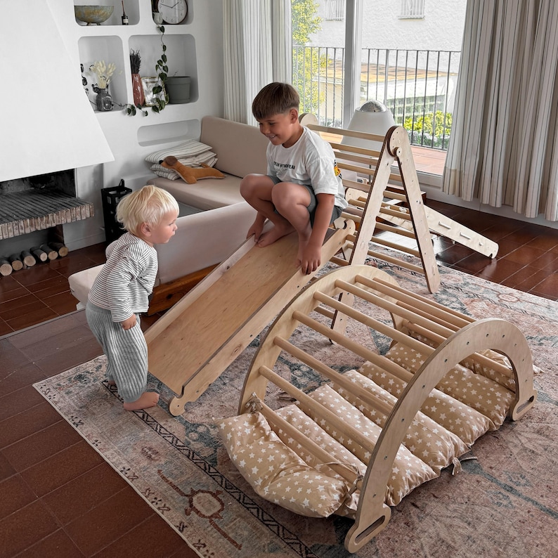 May include: Wooden climbing arch, slide, and climbing triangle set. The arch has a series of wooden rungs and is paired with star-patterned cushions. The slide and triangle are also made of wood. Two children are playing on the set in a living room.
