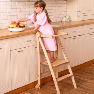 May include: A natural wood step stool with a child standing on it, reaching for food on a countertop. The stool has a platform and a handle for support. The child is wearing a pink outfit. The kitchen has white cabinets and a light-colored countertop.