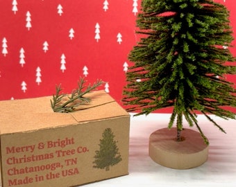 May include: A small green artificial Christmas tree with a brown base, sitting on a white surface in front of a red background with white Christmas tree silhouettes. The tree is packaged in a brown box with the text "Merry & Bright Christmas Tree Co. Chattanooga, TN Made in the USA".