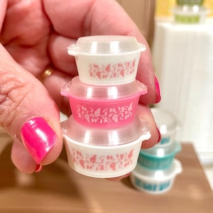 May include: A stack of miniature, vintage-style Pyrex bowls with clear lids. The bowls are white and pink, with a floral pattern. The stack is held in a hand with bright pink nail polish. Other bowls are visible in the background.