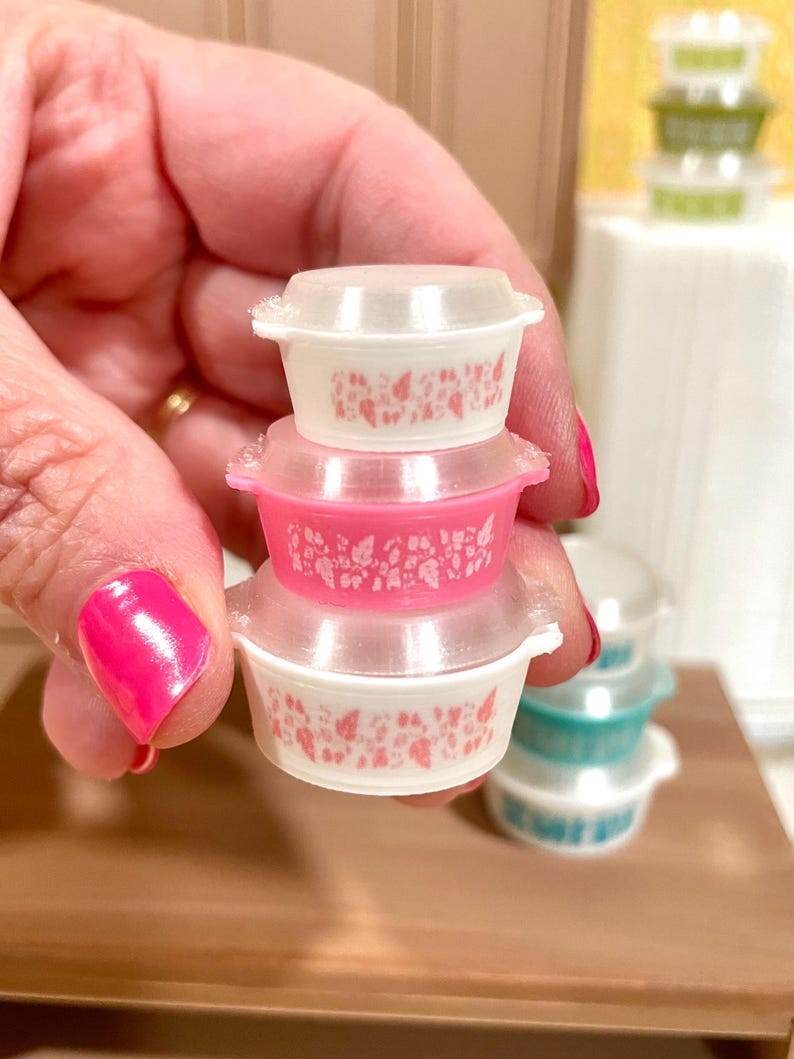 May include: A stack of miniature, vintage-style Pyrex bowls with clear lids. The bowls are white and pink, with a floral pattern. The stack is held in a hand with bright pink nail polish. Other bowls are visible in the background.