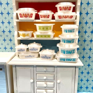 May include: A white miniature cabinet displays a collection of vintage-style casserole dishes. The dishes feature various floral and geometric patterns in red, yellow, green, and blue. The cabinet has a red and yellow interior.