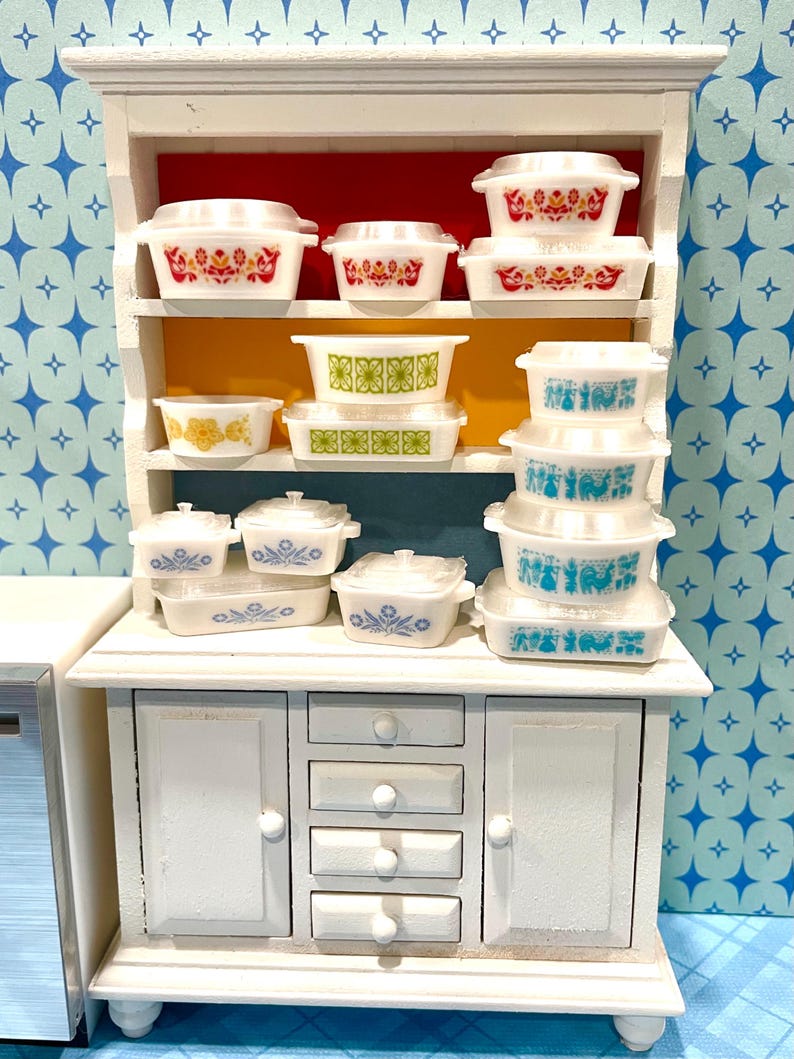 May include: A white miniature cabinet displays a collection of vintage-style casserole dishes. The dishes feature various floral and geometric patterns in red, yellow, green, and blue. The cabinet has a red and yellow interior.