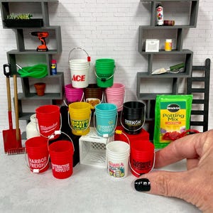 May include: A collection of miniature plastic buckets in various colors, each featuring different brand logos such as Ace, Milwaukee, and Harbor Freight. The buckets are displayed alongside miniature shelving units, tools, and a bag of potting mix.