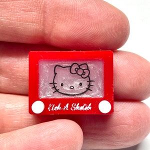 May include: A red Etch A Sketch toy with a white Hello Kitty drawing on the screen. The toy has two white knobs on the bottom.