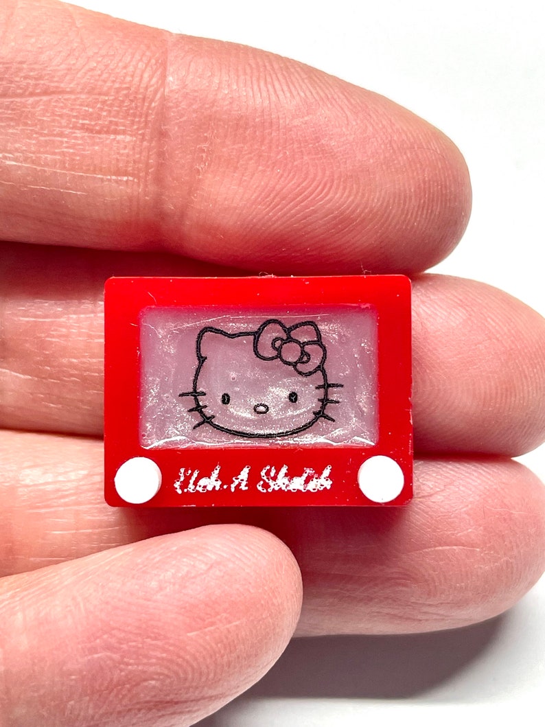May include: A red Etch A Sketch toy with a white Hello Kitty drawing on the screen. The toy has two white knobs on the bottom.