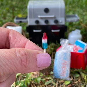 May include: A miniature red, white, and blue popsicle on a stick. The popsicle is being held by a hand with pink nail polish. The popsicle is in focus, while the background is blurred and shows a miniature grill, a cooler, and a bag of ice.