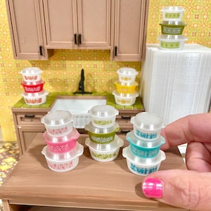 May include: Miniature Pyrex-style bowls in pink, green, and blue with white floral patterns and clear lids. The bowls are stacked on a wooden table in a dollhouse kitchen setting, with a sink and cabinets in the background.