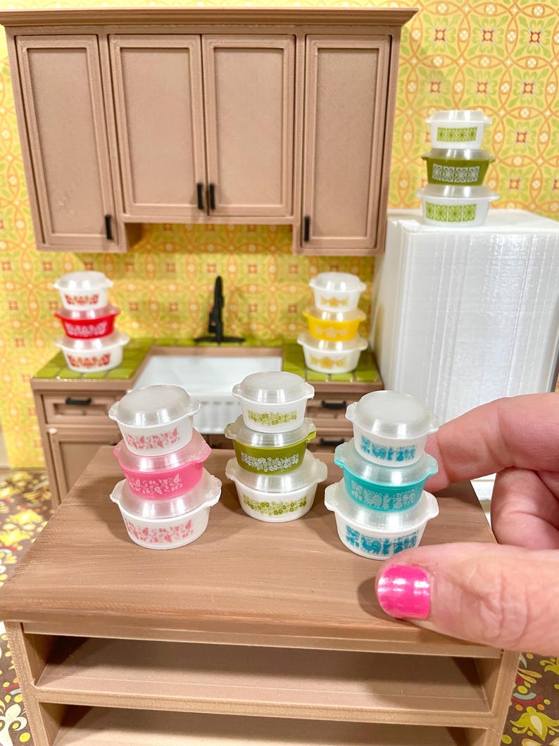 May include: Miniature Pyrex-style bowls in pink, green, and blue with white floral patterns and clear lids. The bowls are stacked on a wooden table in a dollhouse kitchen setting, with a sink and cabinets in the background.