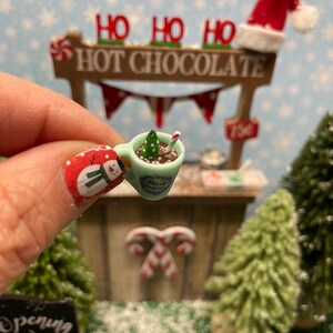 May include: A miniature Christmas hot chocolate stand with a green and white mug filled with hot chocolate, a candy cane, and a small green Christmas tree. The stand has a sign that says "Ho Ho Ho Hot Chocolate".