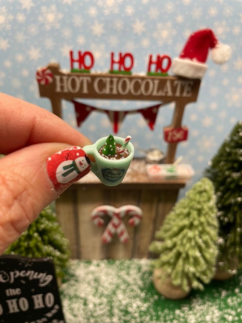May include: A miniature Christmas hot chocolate stand with a green and white mug filled with hot chocolate, a candy cane, and a small green Christmas tree. The stand has a sign that says "Ho Ho Ho Hot Chocolate".