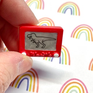 May include: A red plastic toy Etch A Sketch with a black and white drawing of a dinosaur inside. The toy is being held in a hand.