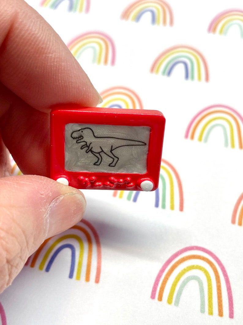 May include: A red plastic toy Etch A Sketch with a black and white drawing of a dinosaur inside. The toy is being held in a hand.