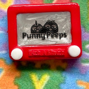 May include: A red plastic toy Etch A Sketch with a silver screen. The screen shows a black and white drawing of three cartoon characters with the text "Punny Peeps" below. The bottom of the toy has the text "Etch A Sketch" in white.
