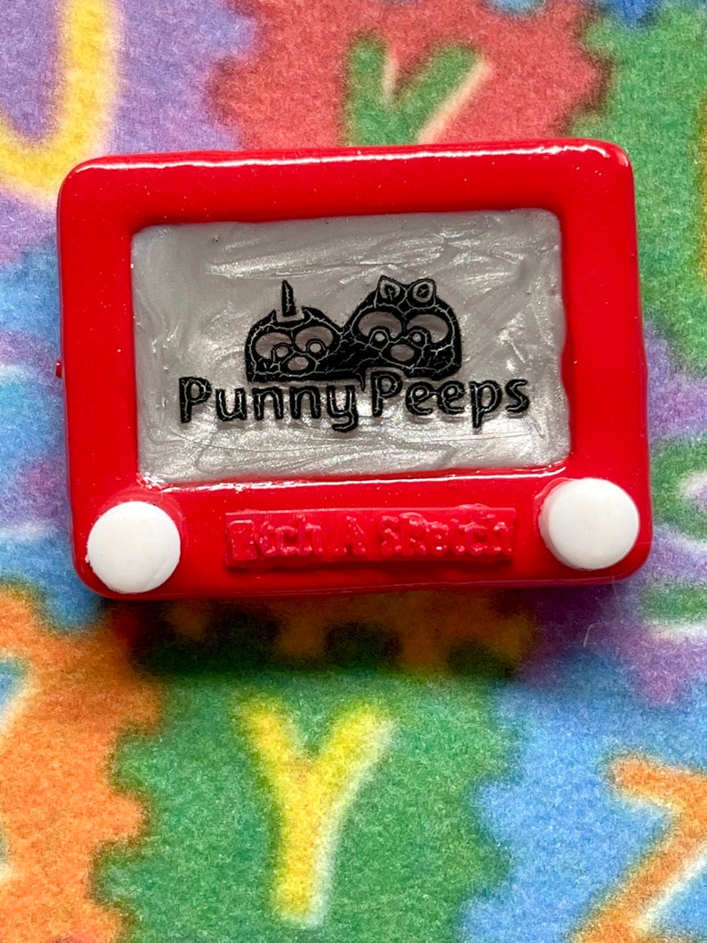 May include: A red plastic toy Etch A Sketch with a silver screen. The screen shows a black and white drawing of three cartoon characters with the text "Punny Peeps" below. The bottom of the toy has the text "Etch A Sketch" in white.