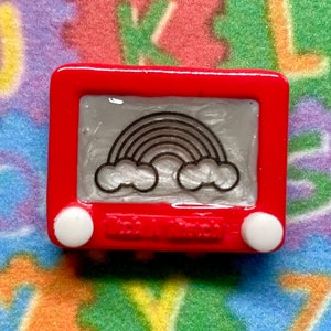 May include: A red plastic toy Etch A Sketch with a rainbow drawing on the screen. The toy has two white knobs on the side.