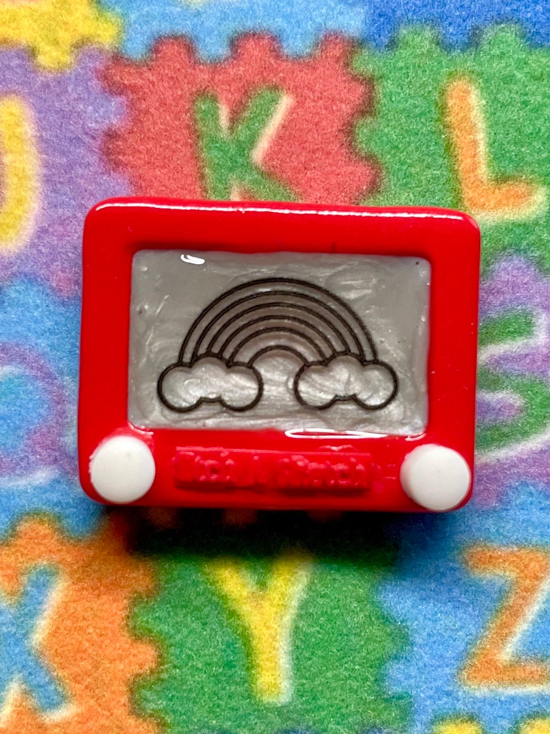 May include: A red plastic toy Etch A Sketch with a rainbow drawing on the screen. The toy has two white knobs on the side.