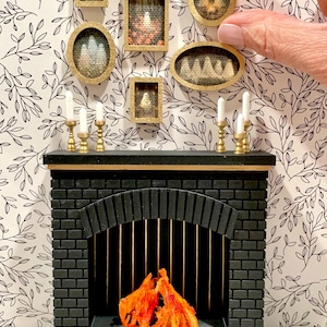 May include: Miniature black fireplace with a brick design, gold candle holders, and a faux fire. Above the fireplace are several small framed pictures with gold frames. The background is a black and white floral wallpaper.