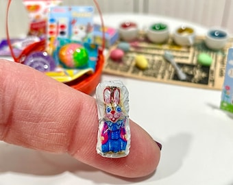 May include: Miniature Easter candy bar featuring a colorful bunny design. The candy bar is wrapped in white paper and held on a white plate. Other miniature Easter-themed items are in the background, including a basket and paint pots.