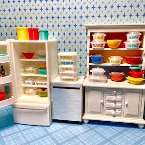 May include: Miniature kitchen scene featuring a white refrigerator, cabinet, and dishwasher. The refrigerator and cabinet are filled with colorful bowls and containers. The scene is set against a blue and white patterned backdrop, creating a retro aesthetic.