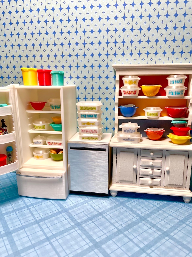 May include: Miniature kitchen scene featuring a white refrigerator, cabinet, and dishwasher. The refrigerator and cabinet are filled with colorful bowls and containers. The scene is set against a blue and white patterned backdrop, creating a retro aesthetic.