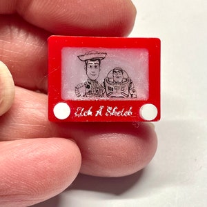 May include: A red plastic Etch A Sketch toy with a black and white drawing of Woody and Buzz Lightyear from the Toy Story movie franchise. The text "Etch A Sketch" is printed on the bottom of the toy.