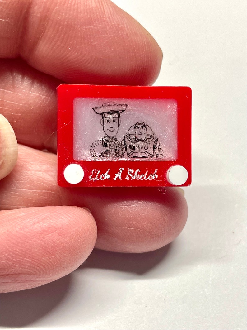 May include: A red plastic Etch A Sketch toy with a black and white drawing of Woody and Buzz Lightyear from the Toy Story movie franchise. The text "Etch A Sketch" is printed on the bottom of the toy.