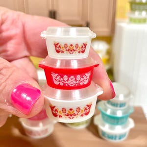 May include: A stack of miniature Pyrex-style bowls with lids. The bowls are white and red, with floral patterns in orange and yellow. Other bowls in green and blue are visible in the background. The bowls are small, likely for a dollhouse or miniature collection.