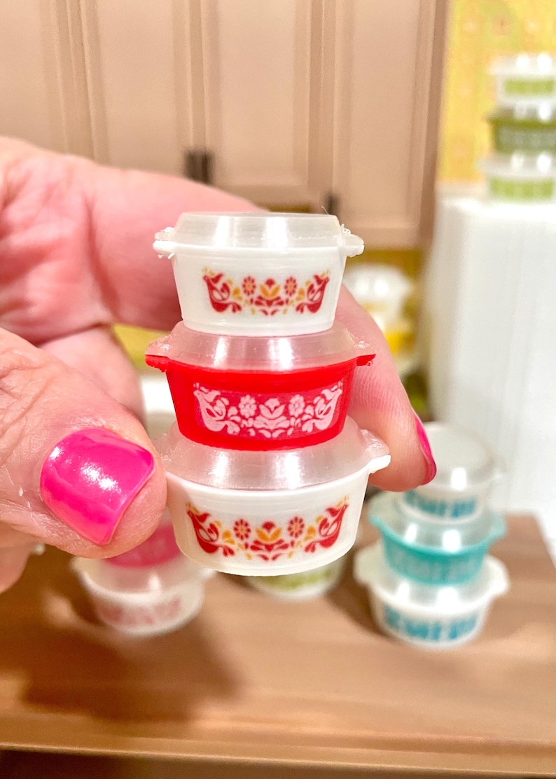 May include: A stack of miniature Pyrex-style bowls with lids. The bowls are white and red, with floral patterns in orange and yellow. Other bowls in green and blue are visible in the background. The bowls are small, likely for a dollhouse or miniature collection.