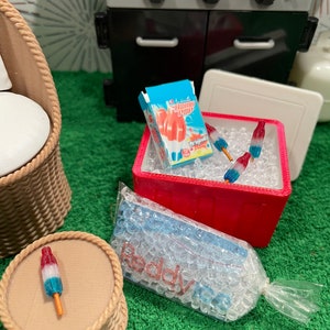 May include: Miniature outdoor barbecue scene with a black grill, a red cooler filled with ice and Bomb Pop popsicles, and a small woven chair. A single popsicle rests on a small round table. A bag of ice is also present.