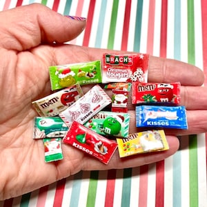 May include: A collection of miniature candy bar wrappers, including M&Ms, Kisses, and Brach's Christmas Nougats. The wrappers are printed with festive holiday designs and feature various colors, including red, green, white, and yellow.