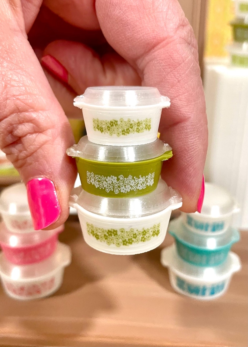 May include: Miniature Pyrex-style bowls in white and green, stacked. The bowls have a floral pattern and clear lids. Other bowls in pink and teal are visible in the background.