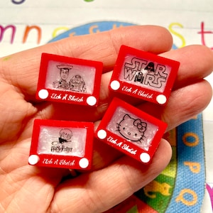 May include: Four red Etch A Sketch toys with different images. The images are of Toy Story characters, Star Wars characters, Hello Kitty, and Harry Potter. Each toy has a white knob on the side.