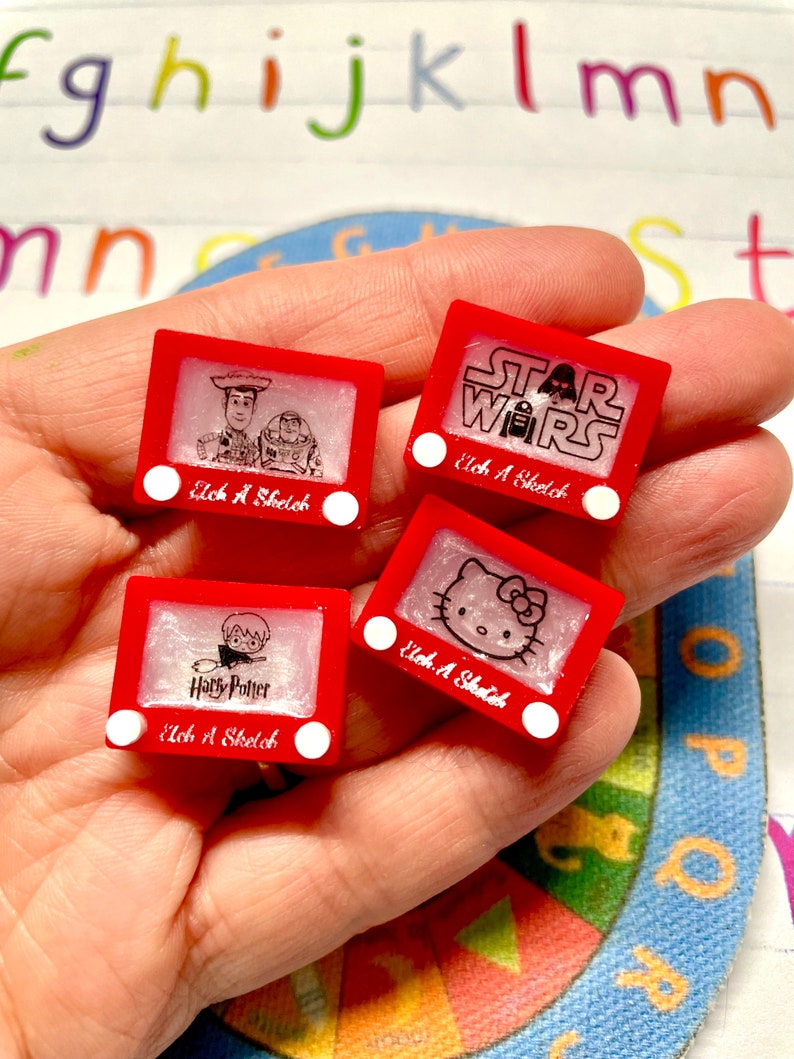 May include: Four red Etch A Sketch toys with different images. The images are of Toy Story characters, Star Wars characters, Hello Kitty, and Harry Potter. Each toy has a white knob on the side.