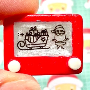 May include: A red plastic toy Etch A Sketch with a white frame and two white knobs. The screen shows a black and white drawing of Santa Claus in a sleigh with presents.