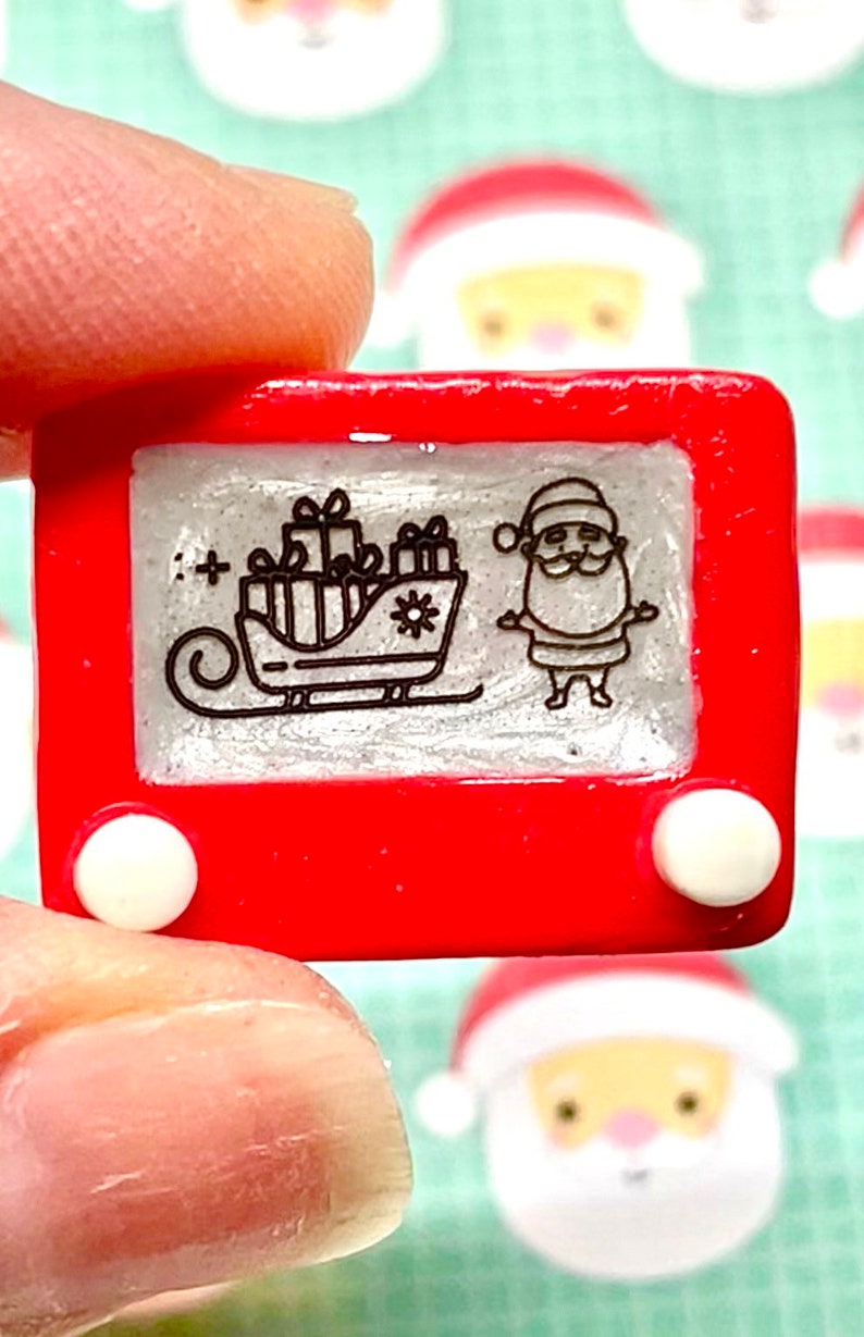 May include: A red plastic toy Etch A Sketch with a white frame and two white knobs. The screen shows a black and white drawing of Santa Claus in a sleigh with presents.