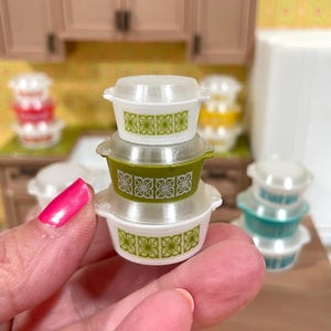 May include: A stack of three miniature white and green casserole dishes with clear lids. Each dish features a repeating green floral pattern. The dishes are held in a person's hand, with other similar dishes in the background.
